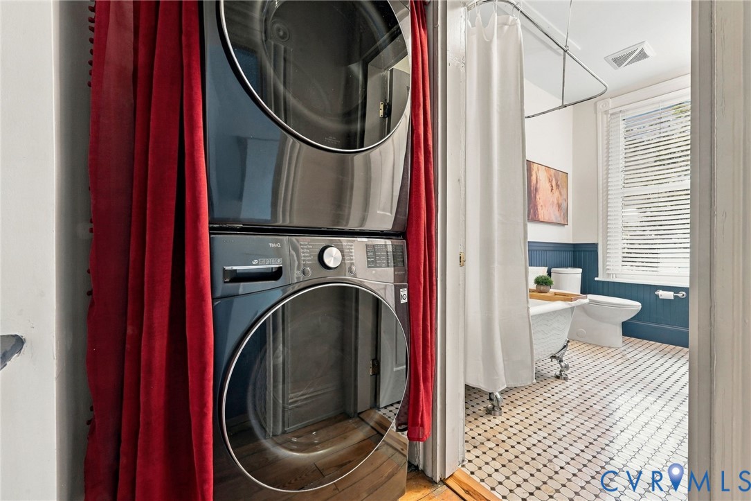 Stackable laundry unit with stainless steel finish, clawfoot tub, dual-tone beadboard wainscoting, geometric tile flooring, and window with shutter blinds