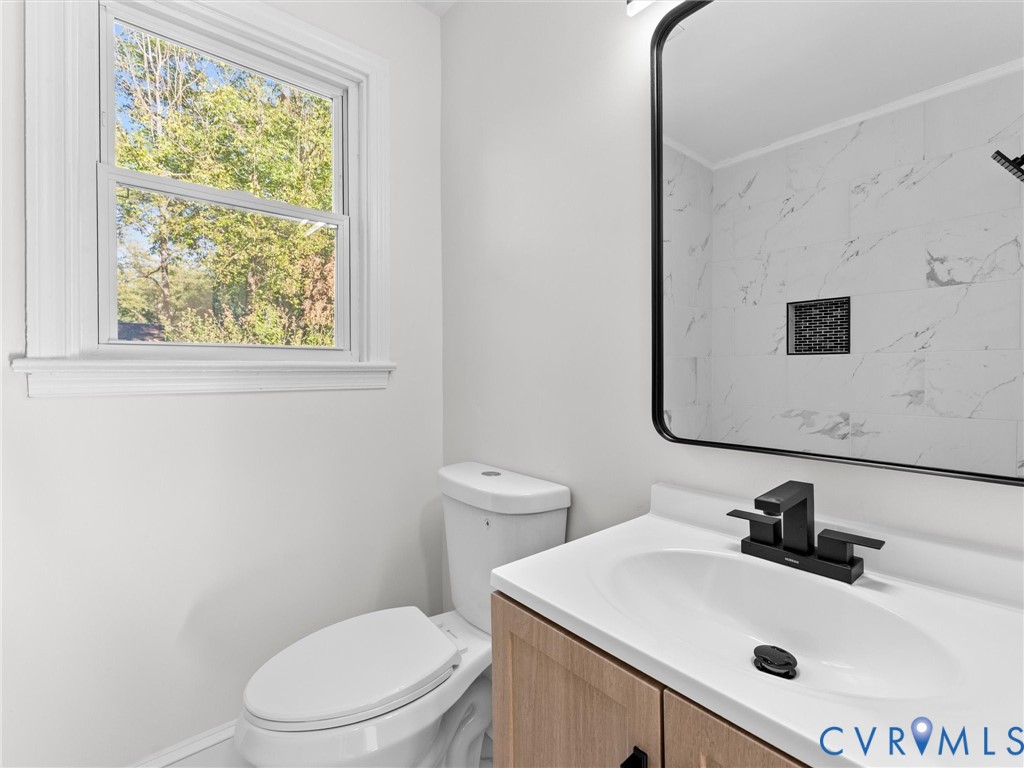 Bathroom featuring a wood-finish vanity with a white countertop, matte black faucet, framed mirror, and tile shower surround with a built-in niche