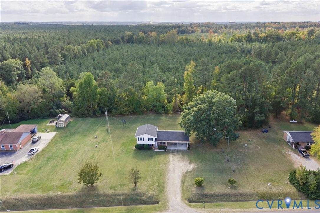 Aerial perspective of a detached residence featuring a covered carport, an expansive lawn, and mature trees, set against a backdrop of dense woodland