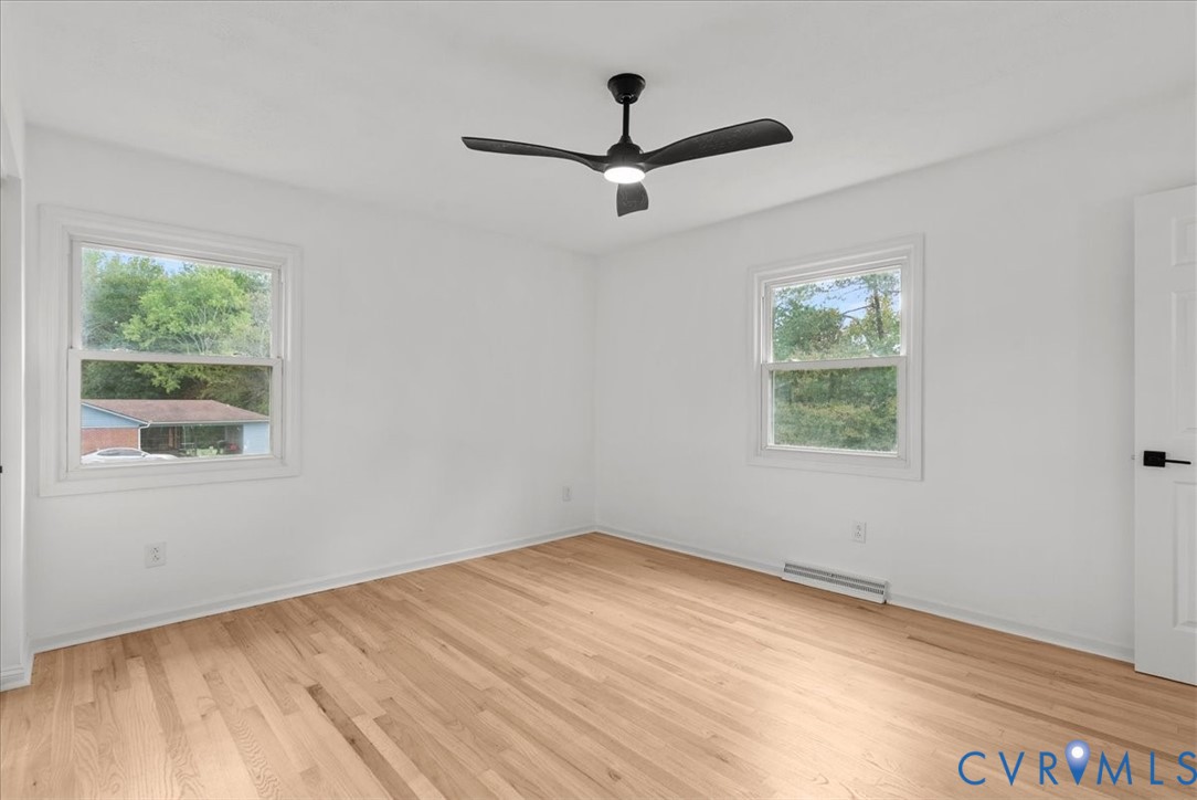 Neutral-toned room featuring light wood-finish flooring, white walls, dual windows, and a matte black ceiling fan with an integrated light fixture