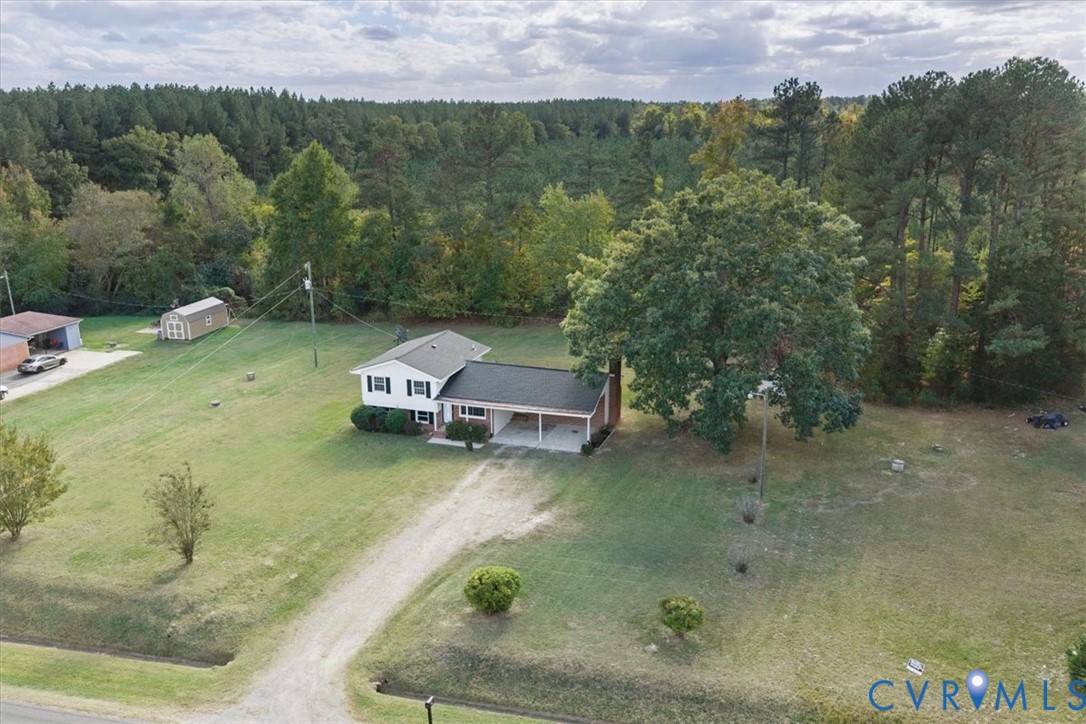Multi-level home featuring an attached carport, light-toned exterior siding, a shingled roof, a detached storage structure, and expansive grounds with mature trees