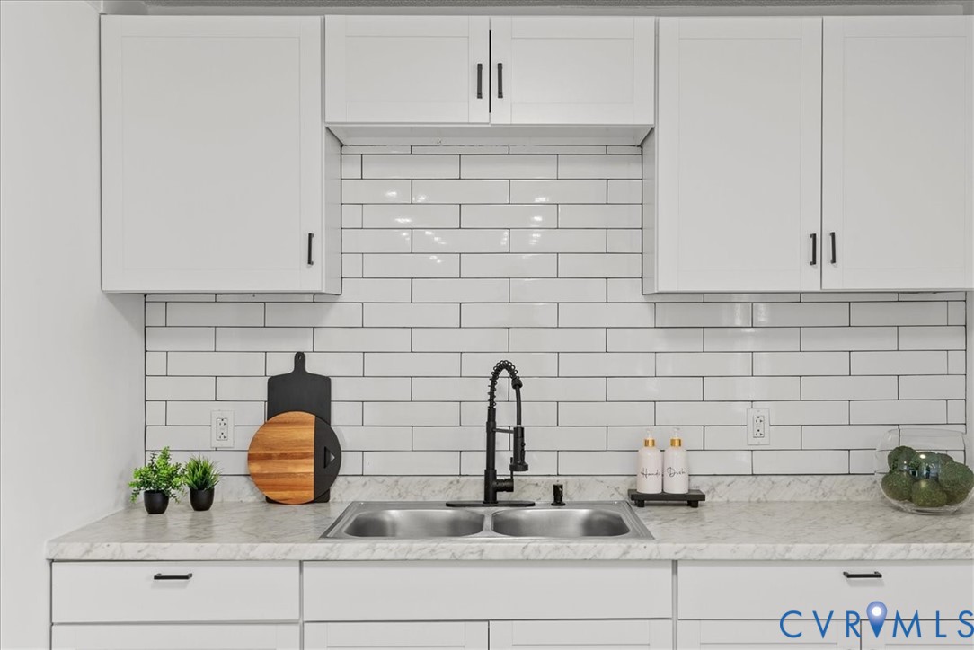 White shaker-style cabinetry featuring matte black hardware, subway tile backsplash, stone-finish countertop, dual-basin stainless steel sink, and high-arc pull-down faucet