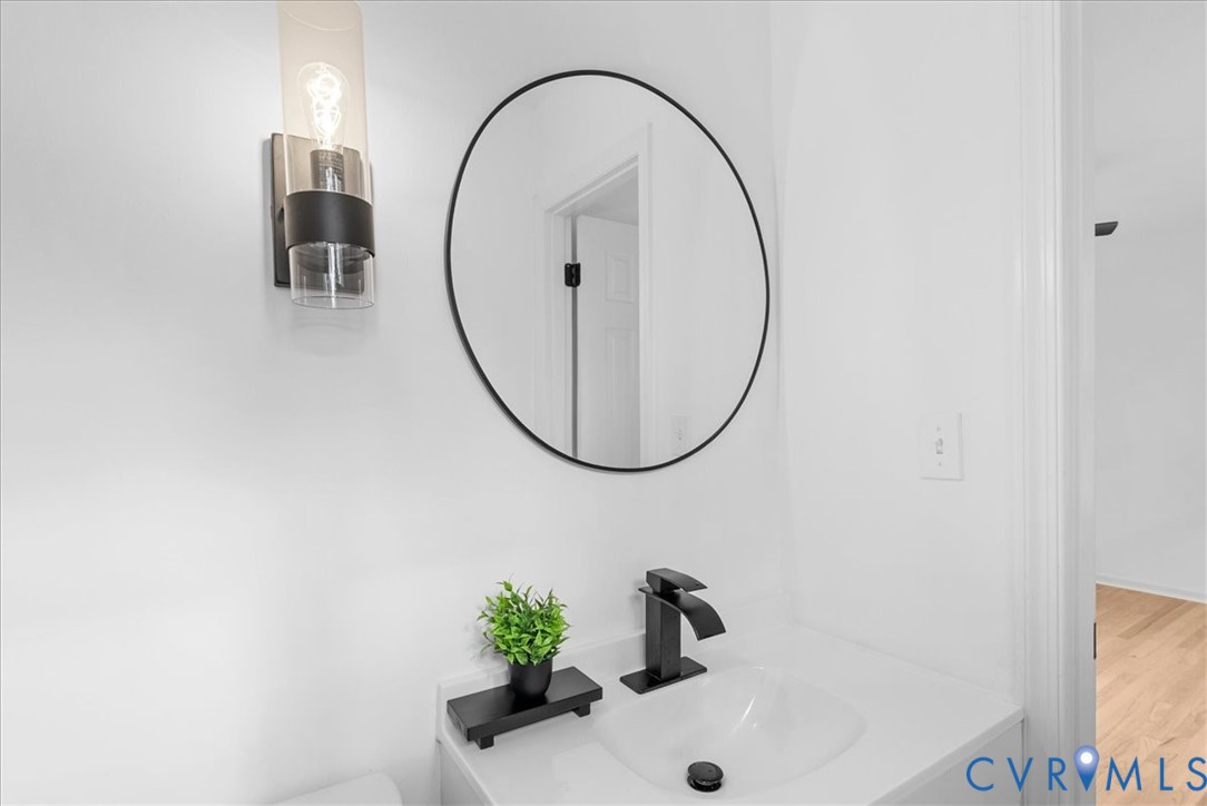 Wall-mounted cylindrical glass and matte black sconce, circular mirror with a slim black frame, integrated vanity countertop with an undermount basin, and matte black single-handle faucet