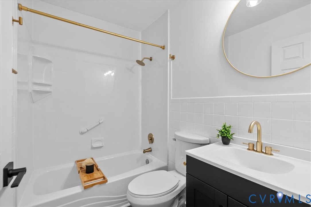 Bathroom featuring a dark-finish vanity with an integrated white countertop, brass-tone hardware, a circular mirror with a matching metallic frame, and a white tub-and-shower enclosure with square wall tile detailing