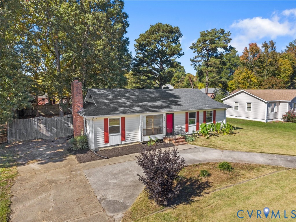 Single-story residence featuring horizontal white siding, a contrasting chimney exterior, red window shutters, a concrete driveway, and mature perimeter trees
