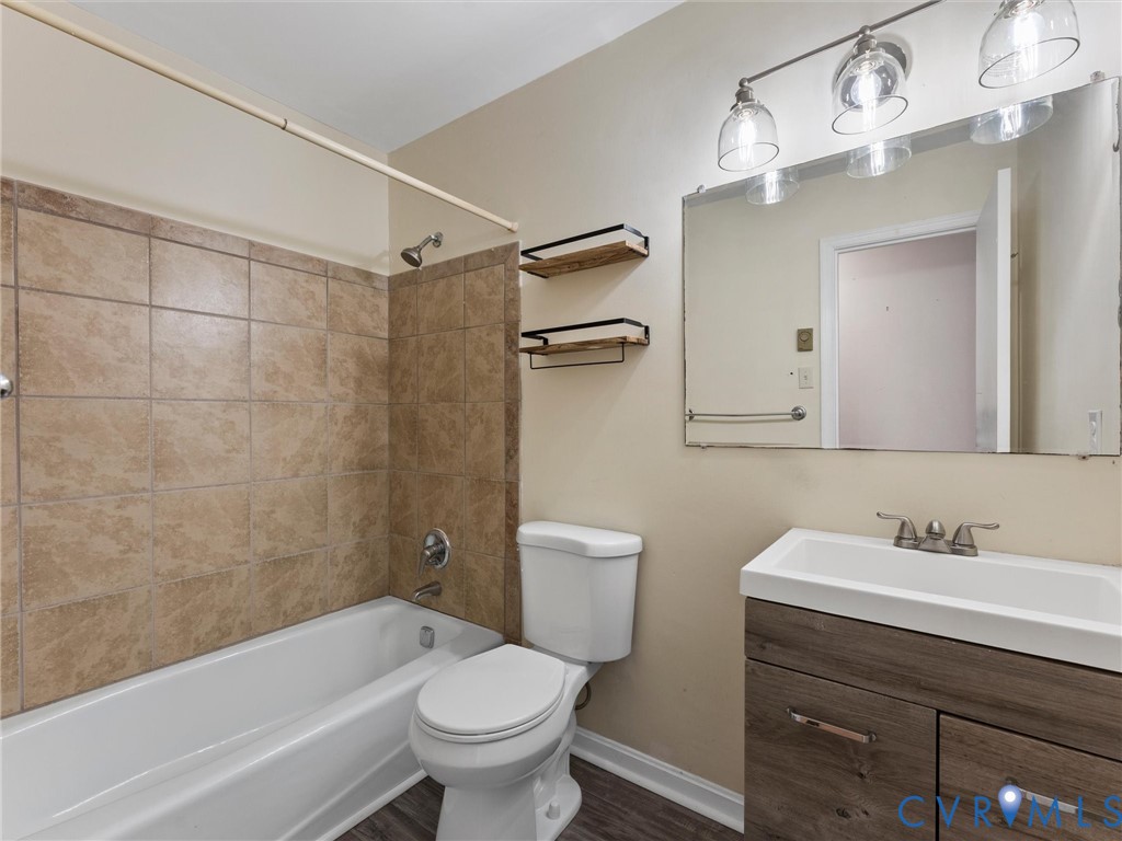 Bathroom featuring a wood-finish vanity with a rectangular sink, wall-mounted light fixture, large vanity mirror, built-in shelving, and a tub enclosure with neutral-toned tiling