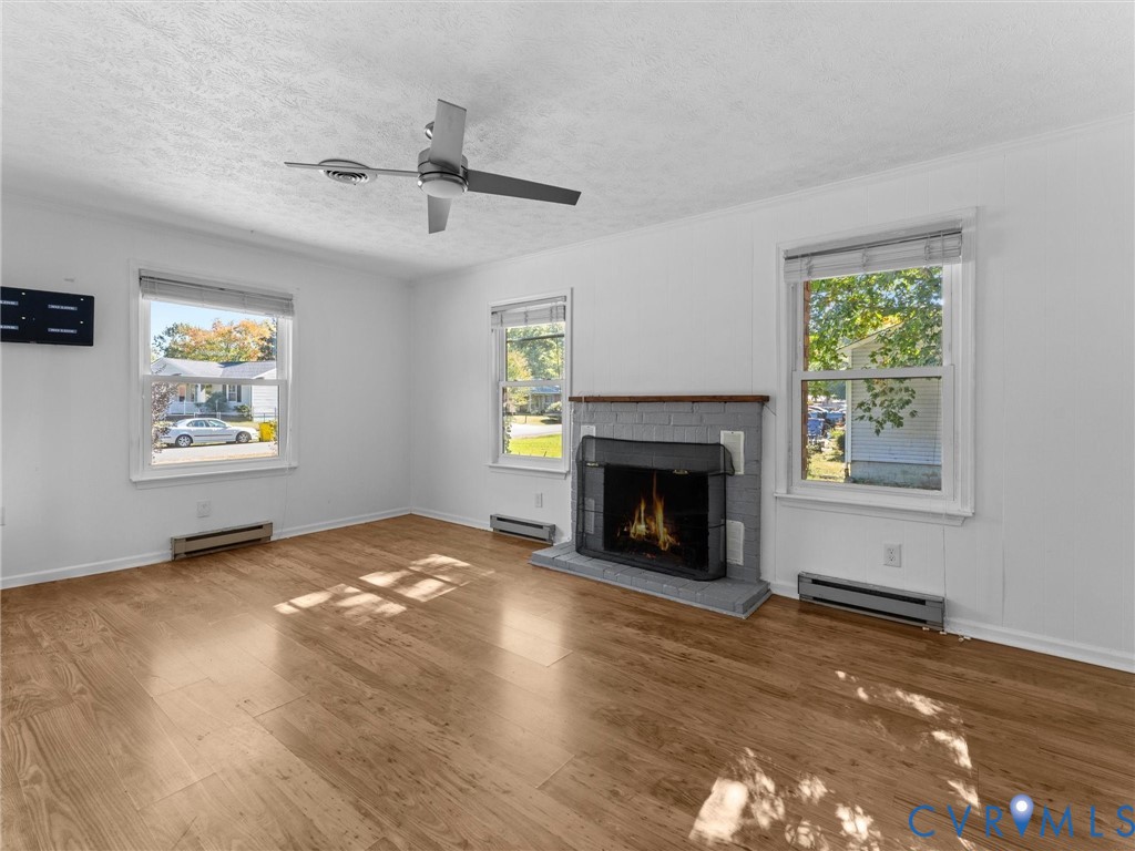 Wood-finish flooring, painted brick fireplace with hearth, metal ceiling fan, and large windows providing natural light