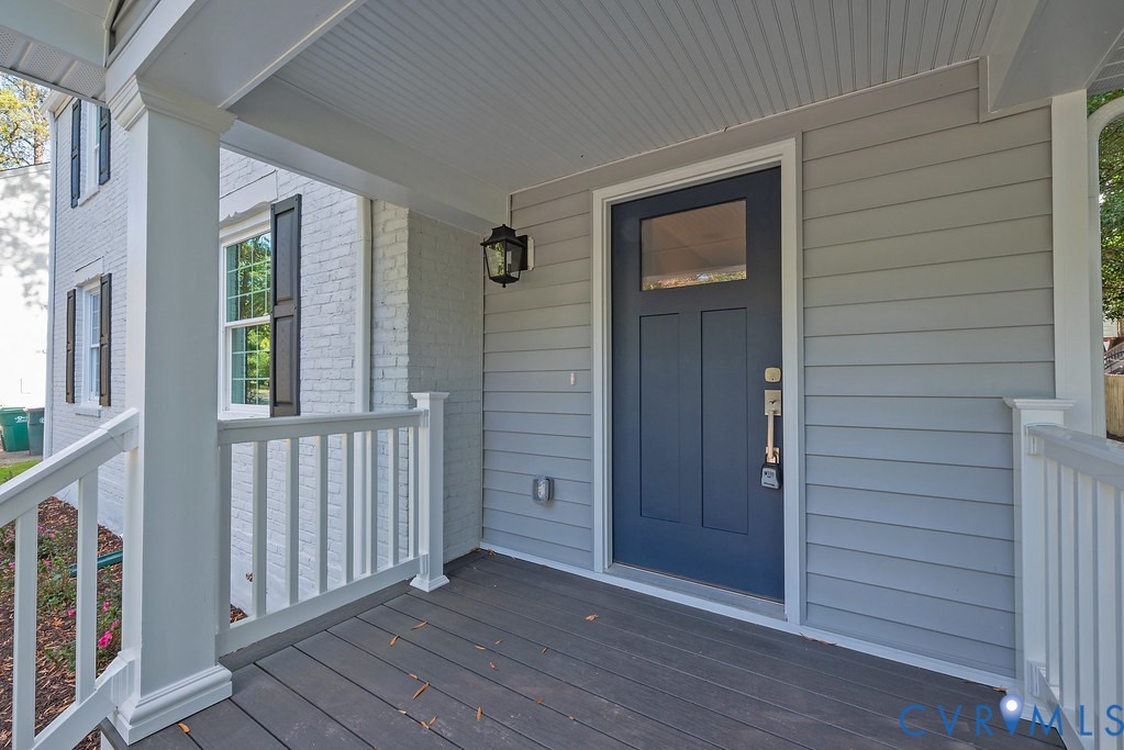 Covered front porch featuring gray wood-finish decking, painted lap siding, a solid-panel door with a single glass pane, white railing, and a wall-mounted lantern light