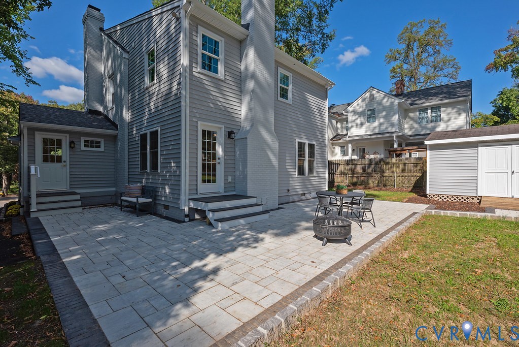 Exterior featuring light gray horizontal siding, a tall painted brick chimney, and a multi-level paver patio with a decorative border