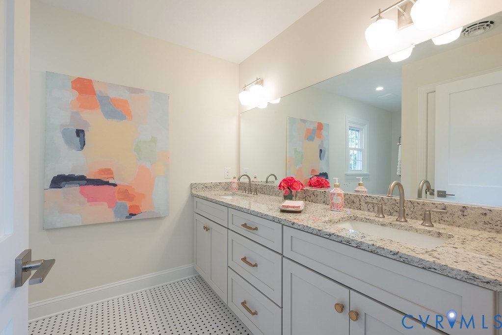 Bathroom featuring a dual-sink vanity with light-toned cabinetry, light-flecked stone countertop, under-mount sinks, brushed metal hardware, and mosaic-style floor tiling