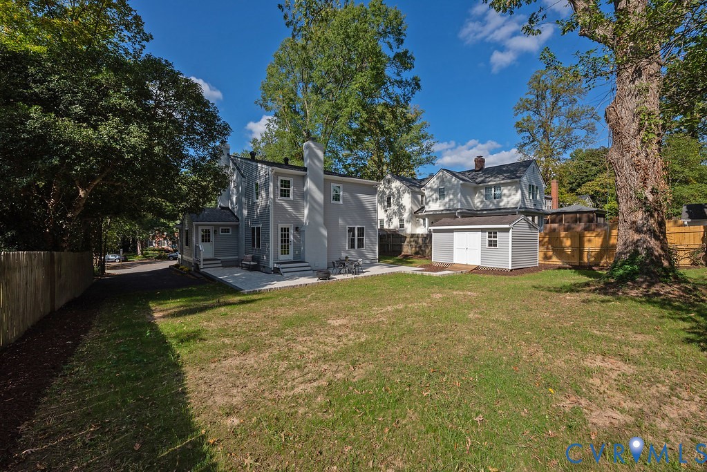 Expansive backyard featuring a manicured lawn, stone patio, detached storage structure, and mature trees, enclosed by wood fencing