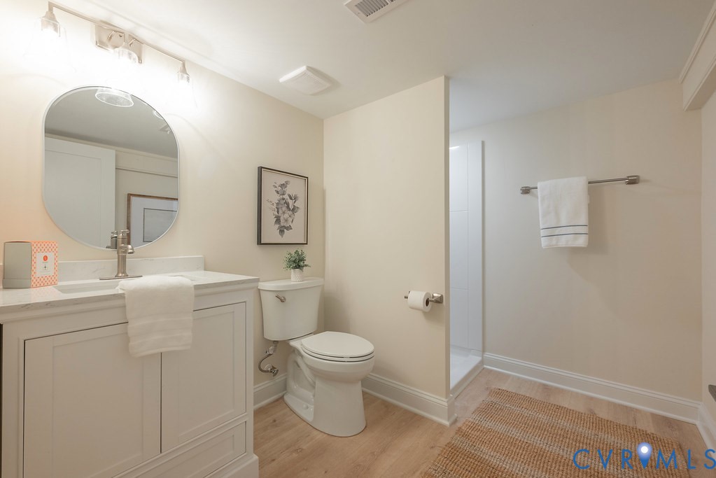 White vanity featuring a stone-composite countertop, undermount sink, arched mirror, and wood-finish flooring, complemented by a walk-in shower with white tile surround
