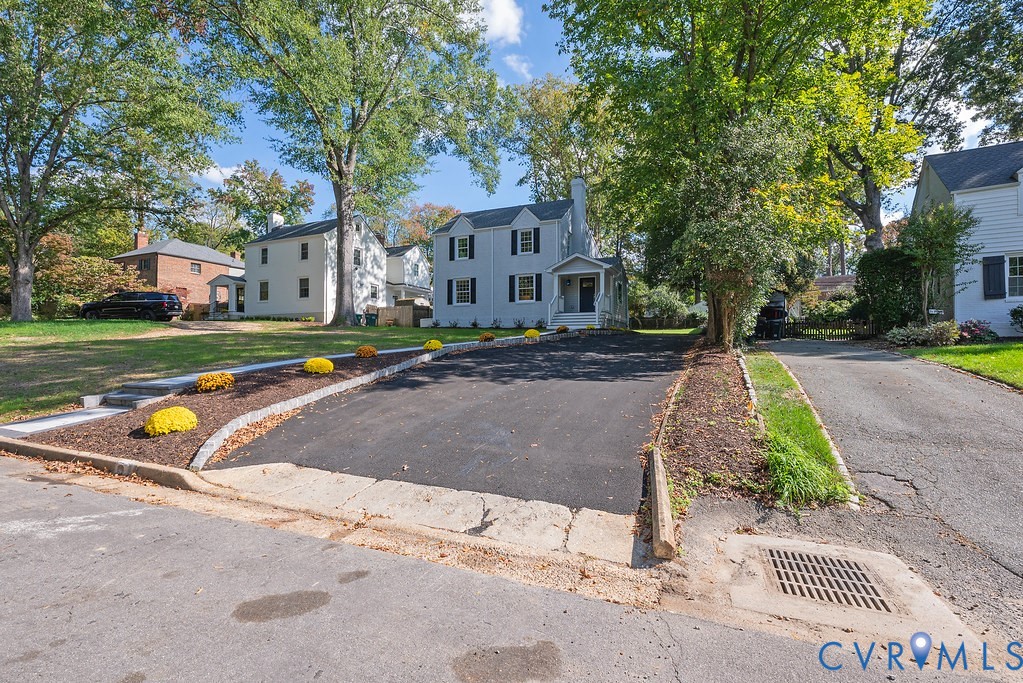 Two-story exterior featuring light-toned siding, dark window shutters, a front chimney, and a paved driveway bordered by stone edging and landscaped mulch beds