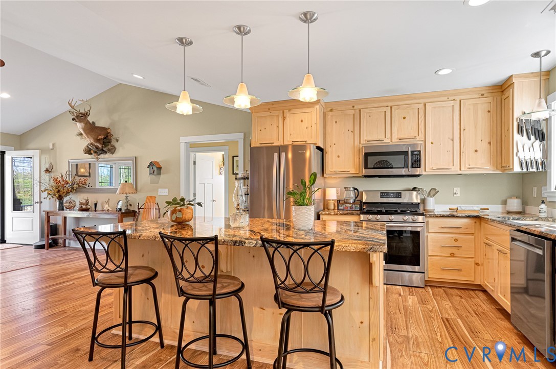 Kitchen featuring light wood cabinetry, stone countertops, stainless steel appliances, wood-finish flooring, and trio of pendant lights over a central island