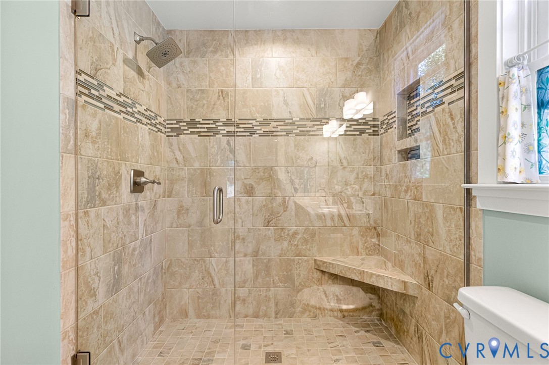 Walk-in shower featuring large-format tile surround, decorative stone mosaic accent border, integrated corner bench, built-in recessed shelf, and glass panel enclosure