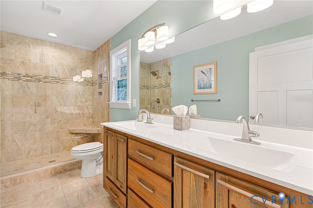 Dual-sink vanity with a solid-surface countertop, walk-in shower with stone tile surround and built-in bench, full-width mirror, and recessed lighting