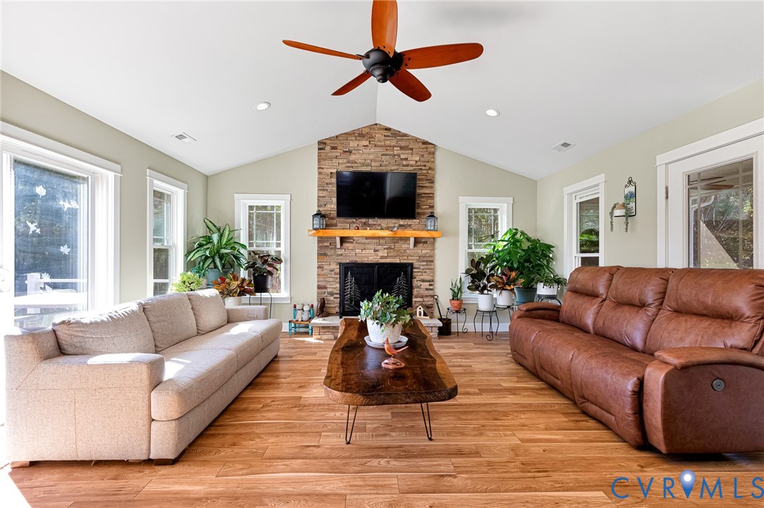 Vaulted ceilings, wood-finish flooring, stone-veneer fireplace with a wood mantel, and a ceiling fan