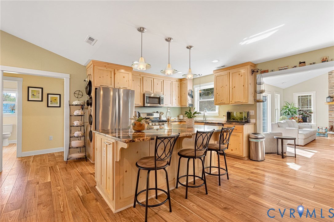 Kitchen featuring natural wood cabinetry, center island with stone-finish countertop, wood-finish flooring, vaulted ceilings, and stainless steel appliances