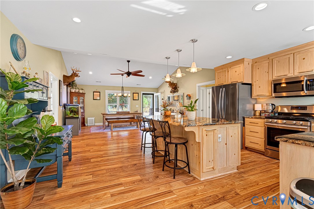 Open-concept kitchen featuring natural wood cabinetry, a central island with a stone-surface countertop, stainless steel appliances, recessed ceiling lighting, and wood-finish flooring
