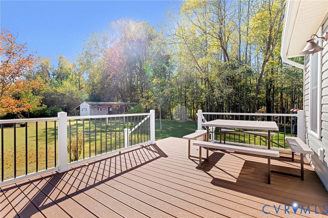 Elevated rear deck featuring composite decking, white posts, black metal balusters, and a wall-mounted exterior light fixture, overlooking a backyard with a detached storage structure and dense tree line