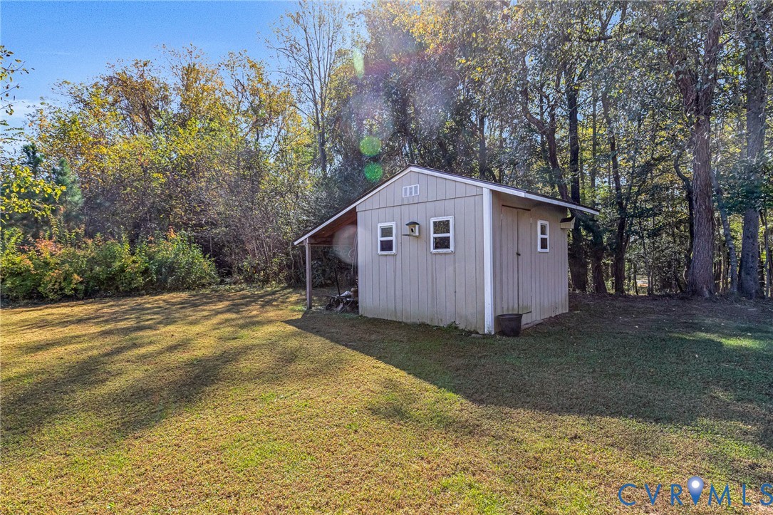 Detached exterior storage structure featuring vertical siding, three windows, and a covered side lean-to, situated on a grass lot with mature perimeter trees