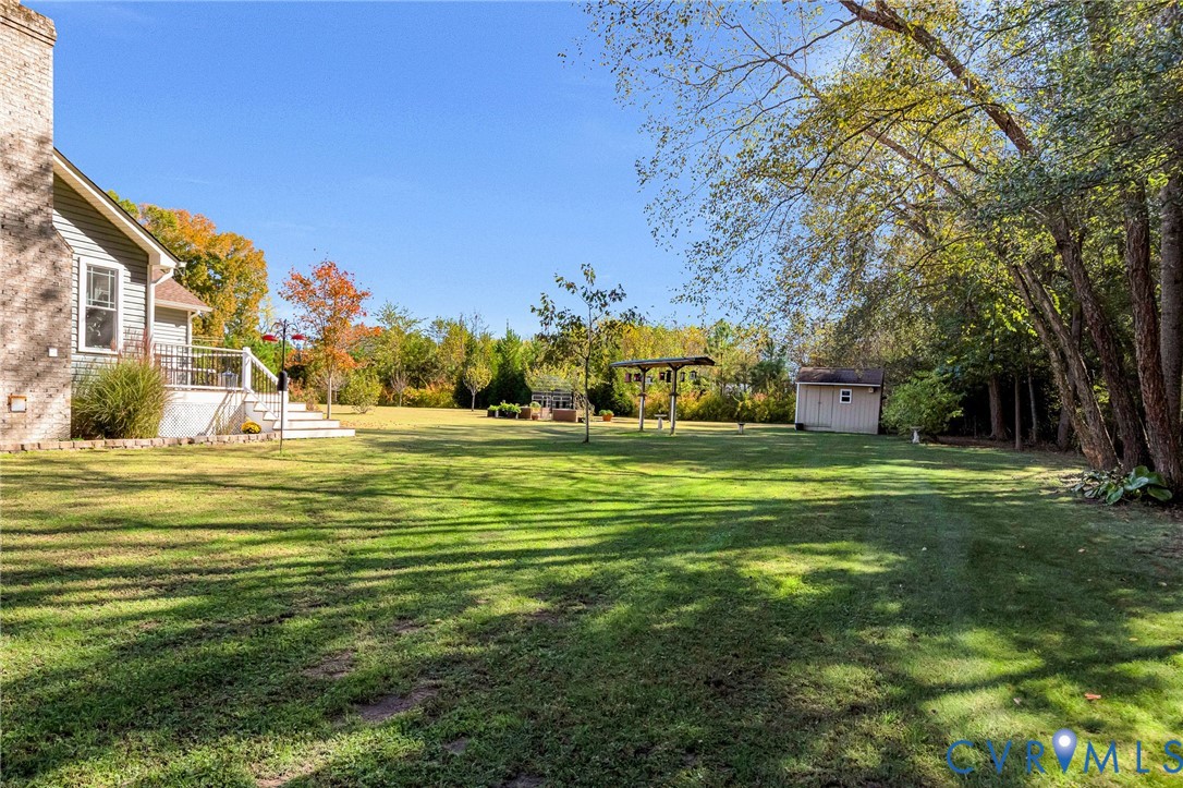 Expansive backyard featuring a brick chimney, raised deck with metal railings, detached storage structure, and mature tree line