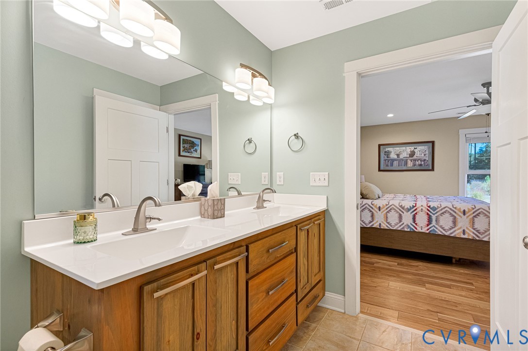 Dual-sink vanity with natural wood-finish cabinetry, solid surface countertop, satin nickel hardware, and large vanity mirror with integrated wall-mounted lighting