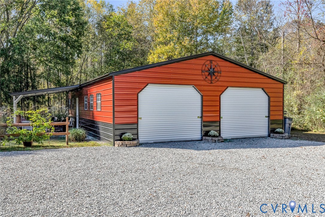 Detached two-car metal garage featuring dual roll-up doors, a side-mounted covered lean-to, and a gravel driveway