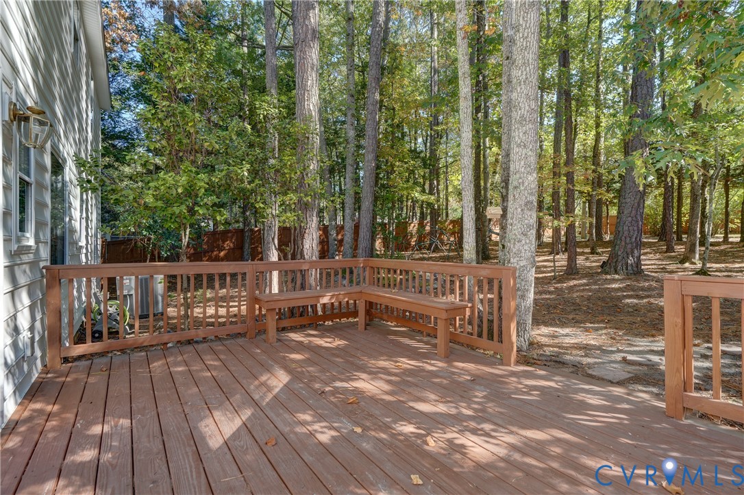 Raised wood-plank deck featuring built-in bench seating, wood railing, exterior wall-mounted light fixture, and wooded surroundings