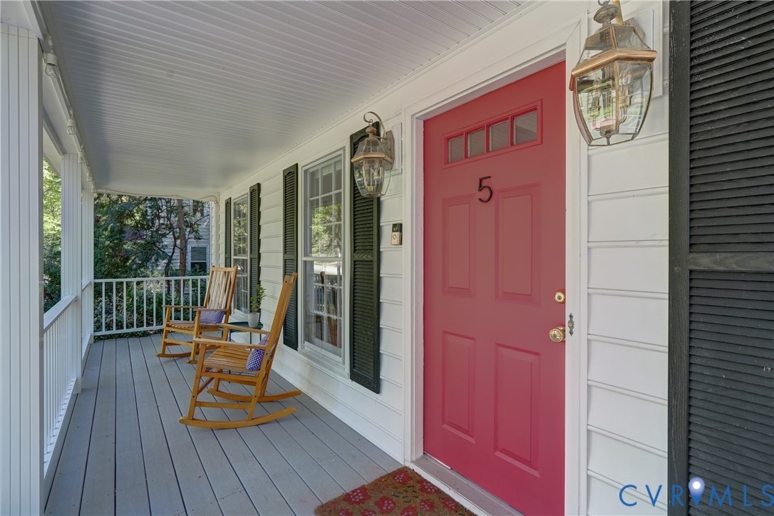 Covered front porch featuring white lap siding, black louvered shutters, a painted entry door with transom glass, and exterior lantern-style sconces
