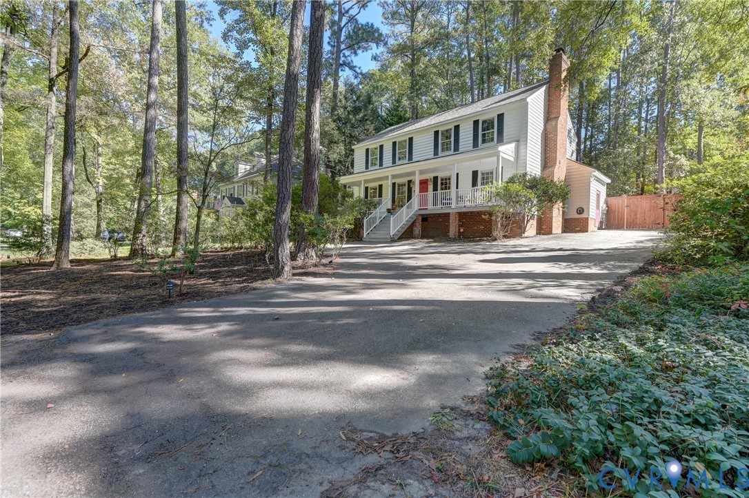 Two-story residence featuring light-colored horizontal siding, a covered front porch with white railing, a side-positioned brick chimney, and a concrete driveway Two-story residence featuring light-colored horizontal siding, a covered front porch with white railing, a side-positioned brick chimney, and a concrete driveway