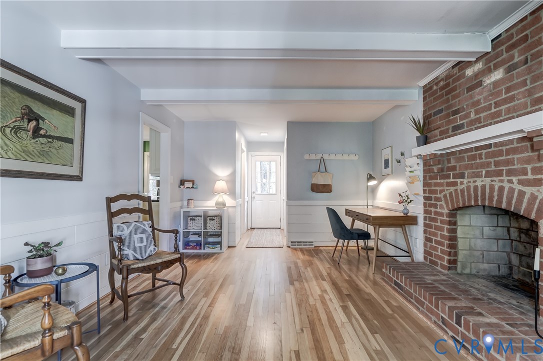 Living area featuring a floor-to-ceiling brick fireplace with a raised brick hearth, exposed ceiling beams, horizontal wainscoting, and wood-finish flooring