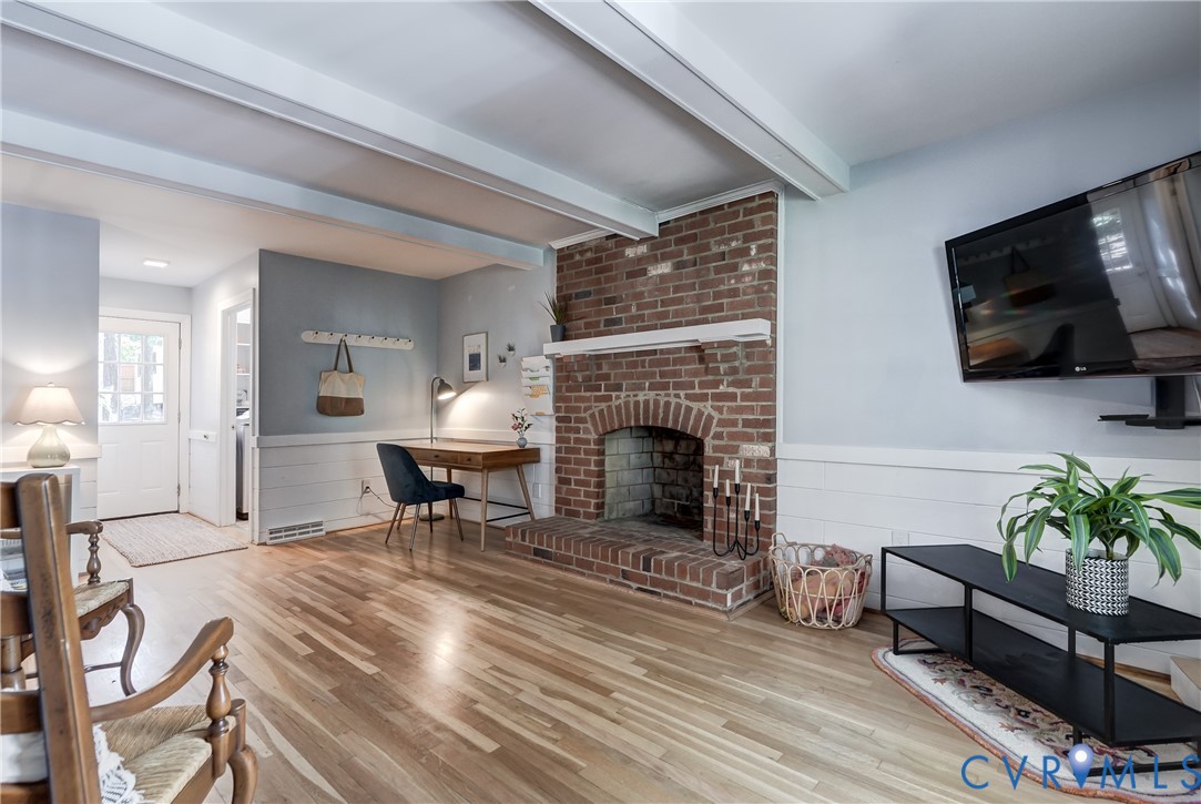 Brick fireplace with a wood mantel, exposed ceiling beams, and horizontal wall paneling over wood-finish flooring