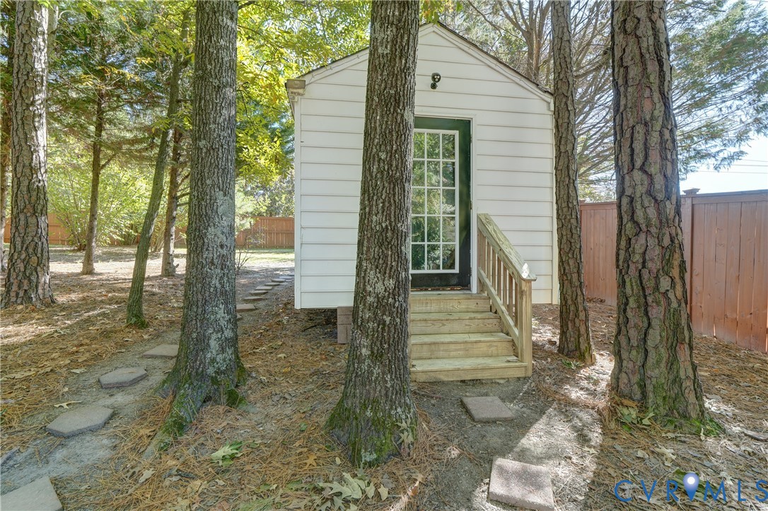 Detached outbuilding featuring white horizontal siding, a glass-paneled entry door, and timber stairs with railings, situated within a wooded lot with stone path pavers and a privacy fence