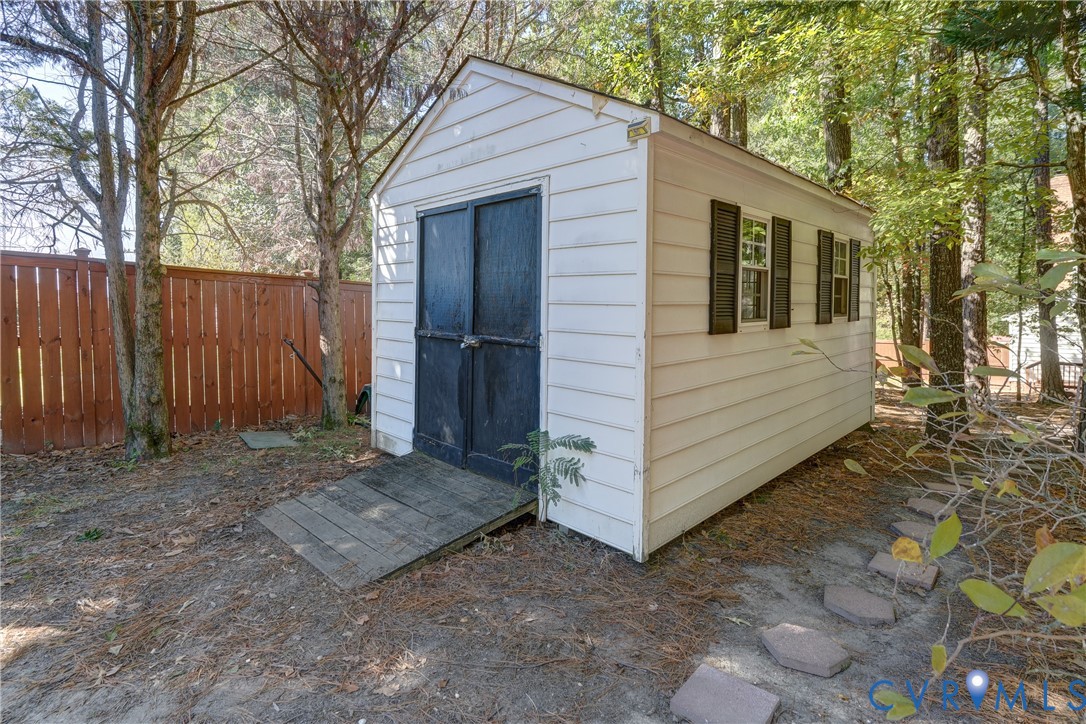 Detached storage structure featuring white horizontal siding, dark shutter-accented windows, double doors with an entry ramp, and a wood-slat perimeter fence