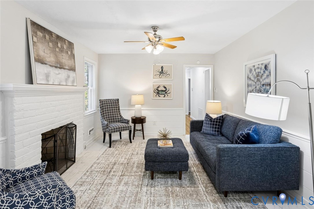 White painted brick fireplace, light-toned wood-finish flooring, half-wall wainscoting, and a multi-blade ceiling fan with integrated lighting