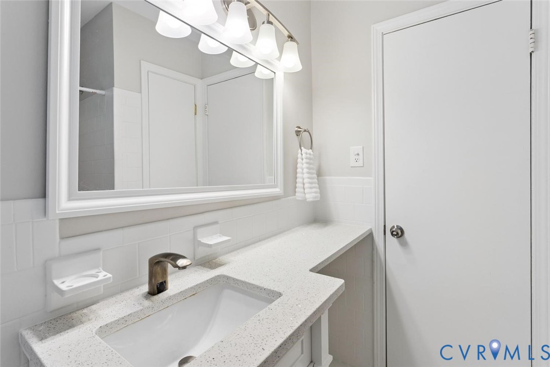 Bathroom vanity featuring a quartz-composite countertop, integrated undermount sink, brushed-metal faucet, white subway tile backsplash, and a framed wall mirror with integrated multi-bulb lighting