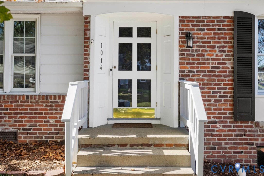 Exterior entrance featuring a brick facade, white multi-pane glass entry door with a brass kick plate, shuttered windows, and concrete steps with white handrails