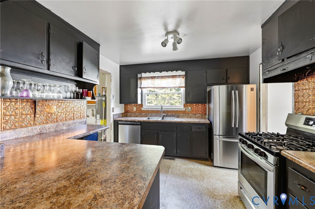 Kitchen featuring matte-finish cabinetry, textured metallic backsplash tiles, stone-pattern countertops, double-basin sink, and stainless steel appliances