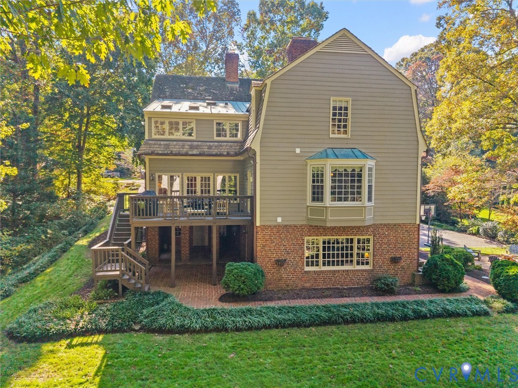 Multi-level exterior featuring a gambrel roofline, brick and siding facade, elevated wood deck with stairs, lower-level brick patio, and bay windows