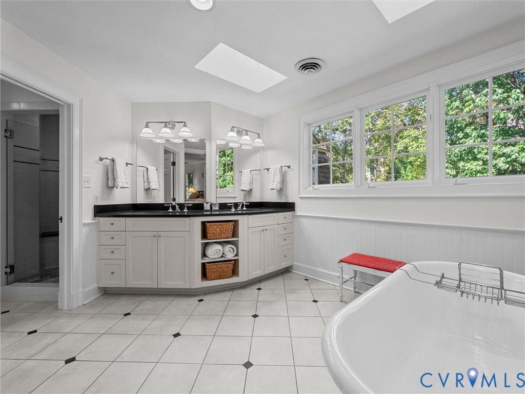 Dual-sink vanity with dark stone countertop, white custom cabinetry, freestanding soaking tub, skylights, and ceramic tile flooring with decorative inserts