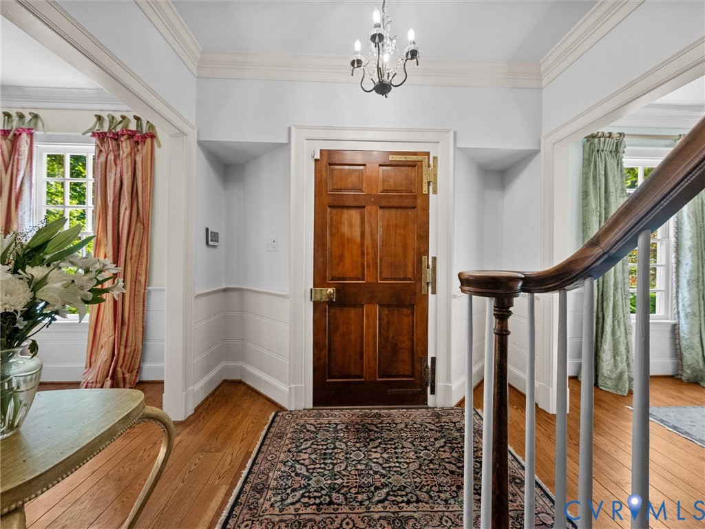 Entryway featuring a six-panel wood door with brass hardware, wood-finish flooring, crown molding, wainscoting, and a multi-light chandelier