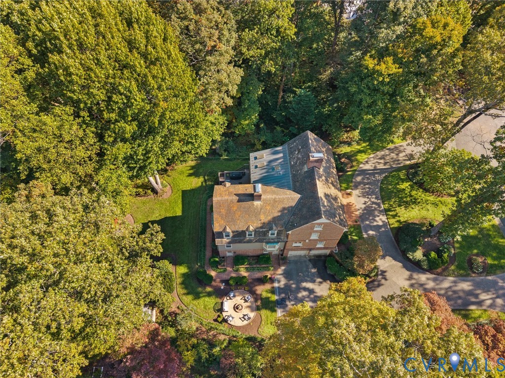 Aerial perspective of a multi-story residence featuring a brick exterior, shingled roof, paved driveway, brick hardscaping, and surrounding mature trees