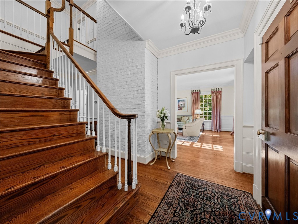 Foyer featuring a hardwood staircase with a wood handrail, white-painted brick detailing, crown molding, and a suspended chandelier