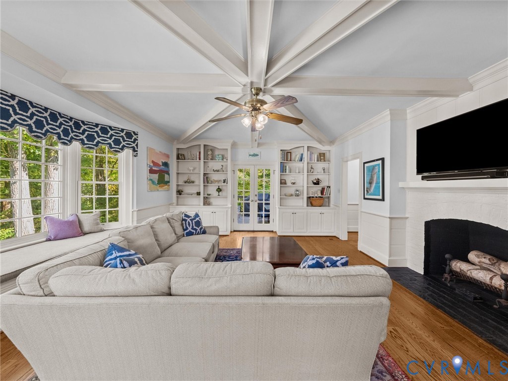 Vaulted ceiling with exposed beams, fireplace with painted brick surround, built-in cabinetry, wood-finish flooring, and multi-pane windows