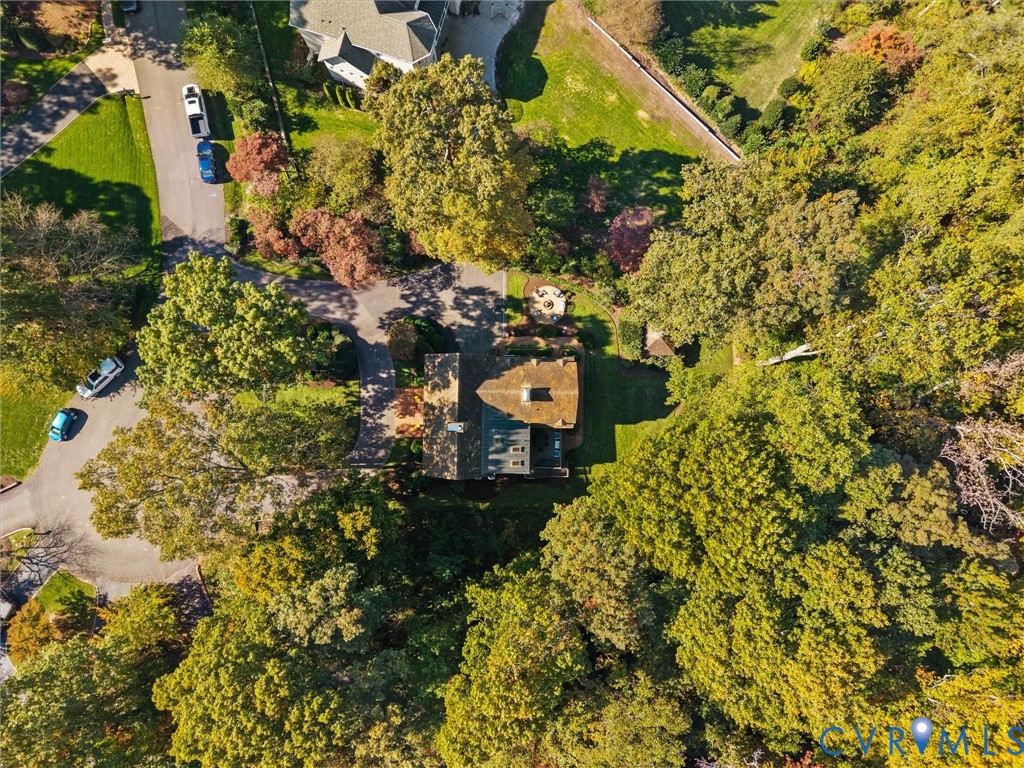 Aerial perspective of a multi-level structure featuring a shingled roof, asphalt driveway, circular stone patio, and extensive mature tree canopy