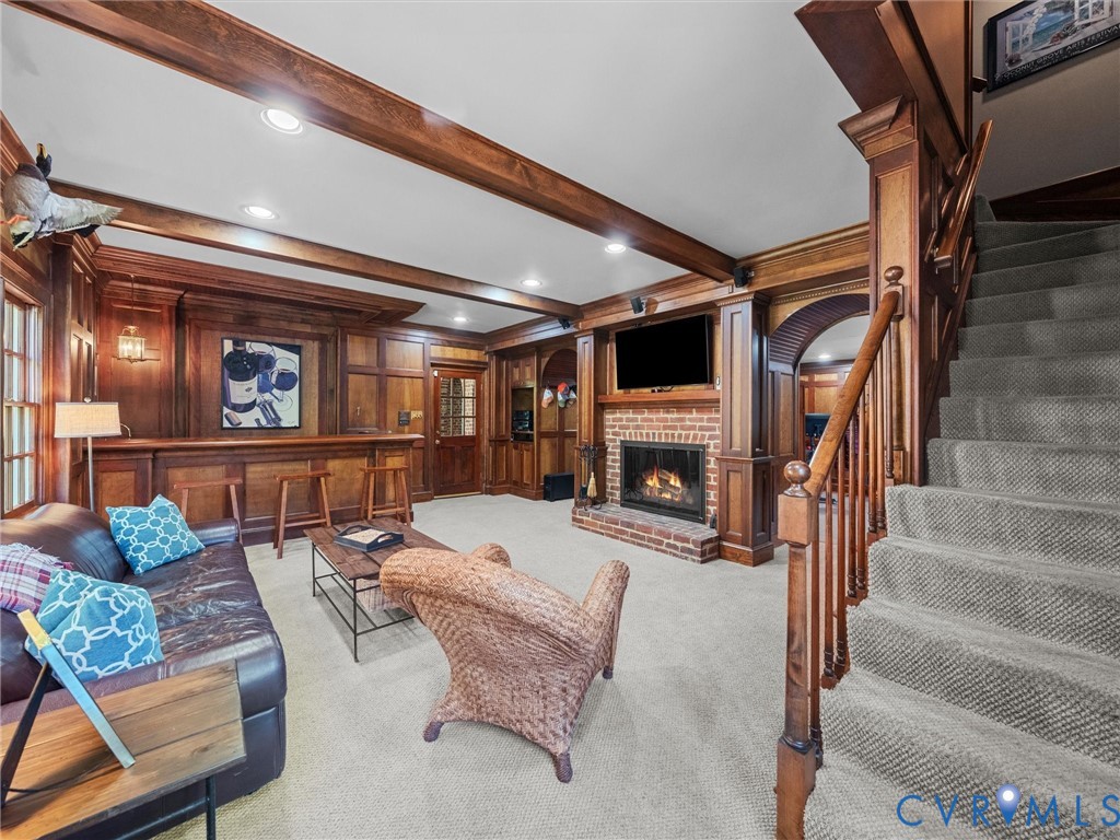 Wood-paneled walls with decorative wainscoting, exposed ceiling beams, brick-surround fireplace, custom built-in bar, and staircase featuring a wood handrail