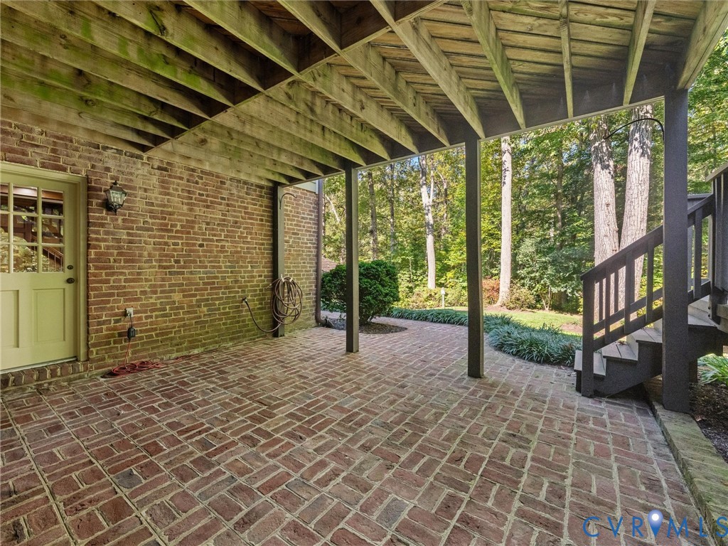 Covered patio featuring a herringbone-patterned brick floor, exposed timber ceiling joists, brick exterior wall, and an attached staircase with wooden railings