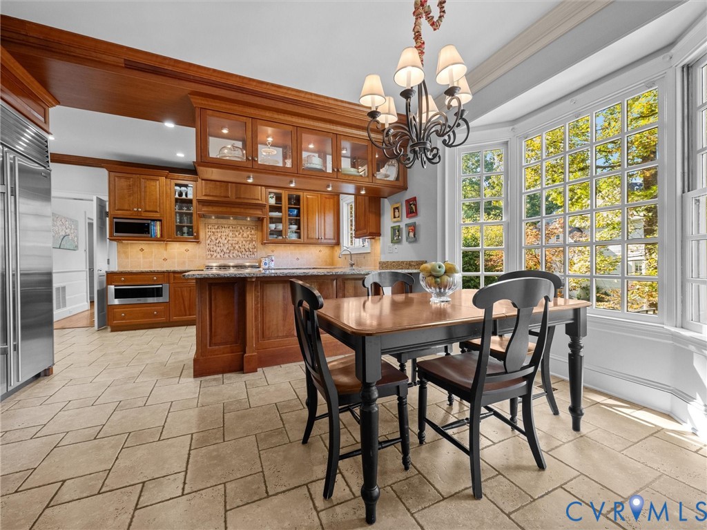 Kitchen featuring custom wood cabinetry with glass-front uppers, stone-surface countertops, stone tile flooring, a large bay window, and a multi-arm chandelier