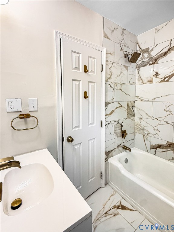 Bathroom featuring large-format decorative wall tile, a standard tub with brass-toned fixtures, a white vanity with an integrated sink, and coordinated brass-toned hardware